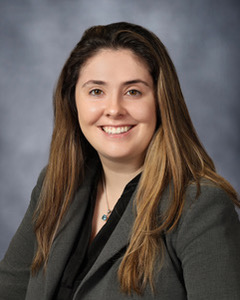 Brooke Brewer 1 A woman with long brown hair smiles, wearing a gray blazer over a black top, set against a plain gray background.