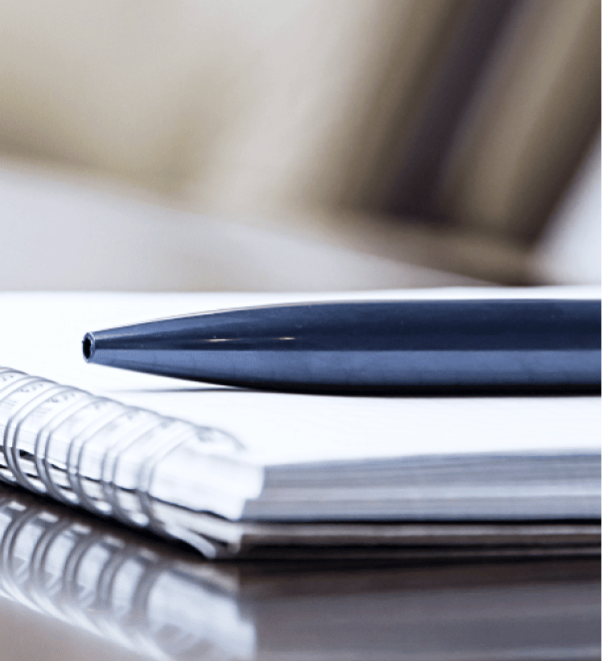 Home 1 Close-up of a black pen resting on an opened spiral notebook, placed on a glossy surface.