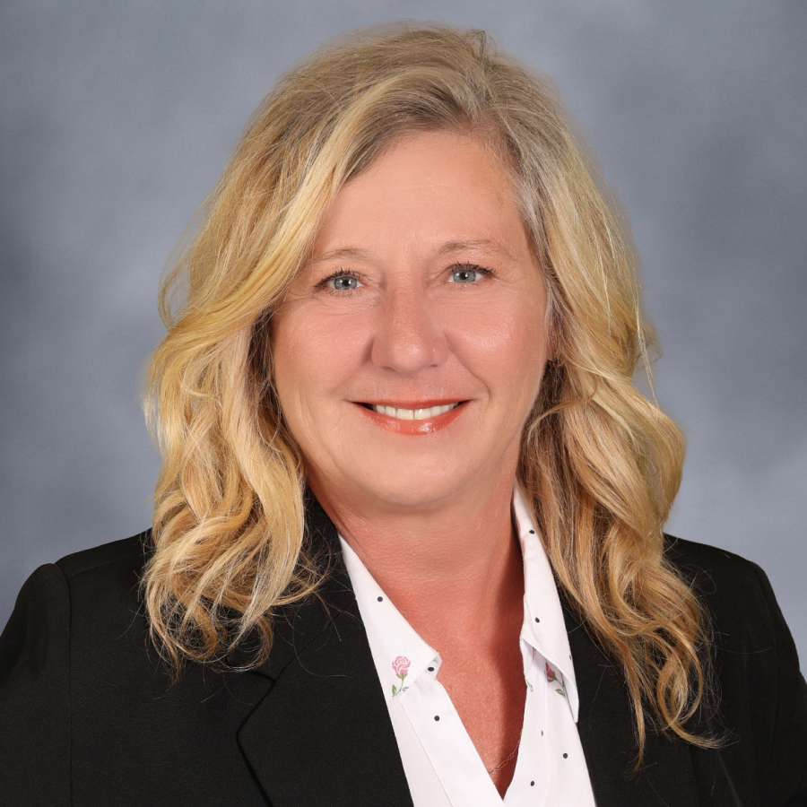 Tracie Zerr 1 A woman with wavy blonde hair, wearing a black blazer over a white blouse, smiles at the camera against a gray background.