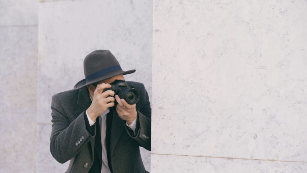 Why Hire a Private Investigator for Your California Divorce 1 A private investigator wearing a hat and coat stands behind a marble wall, holding a camera up to their face and taking a photo.
