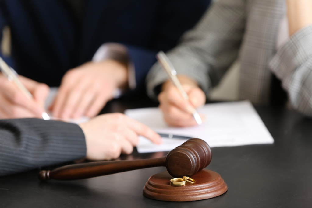 A judge's gavel and two wedding rings on a table hint at hidden assets, as people in suits sign documents in the background during California divorces.