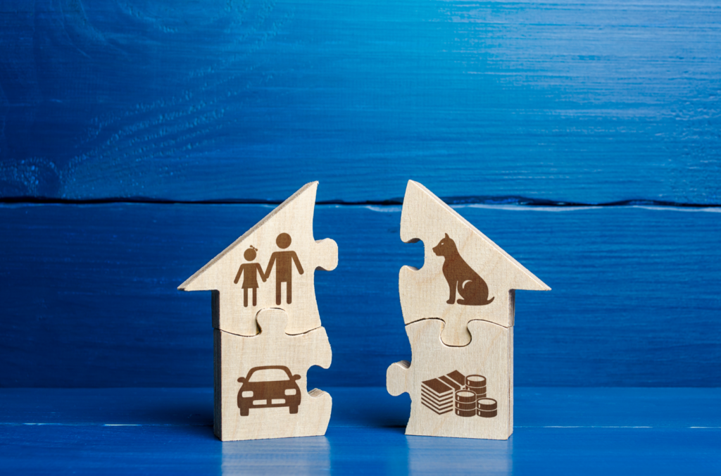 Two wooden house-shaped puzzle pieces against a blue background, one with icons of people and a car, the other with icons of a dog and stacked coins—illustrating fair and equal division in a California divorce.