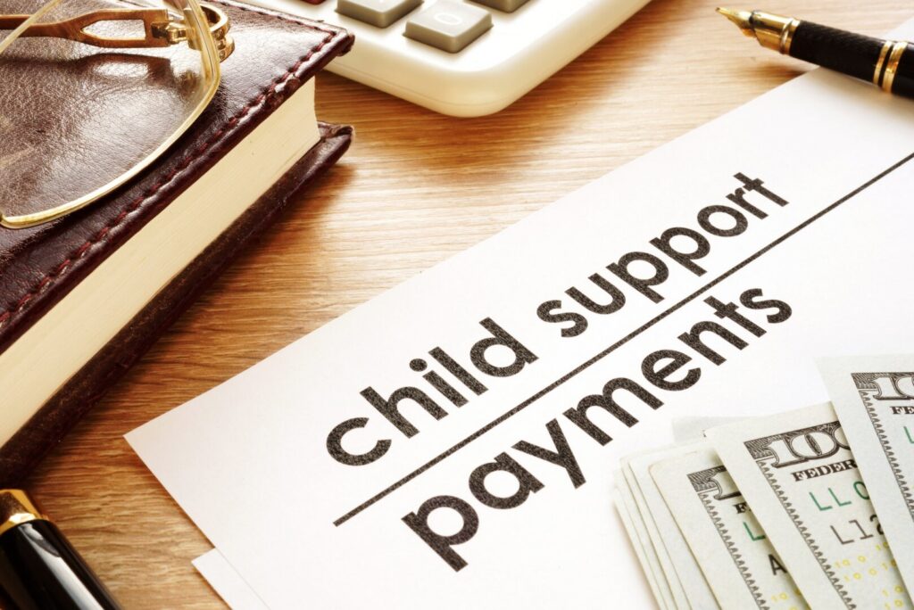 A document labeled "child support payments" sits on a desk with cash, a pen, a calculator, and a closed leather book nearby—an organized scene reflecting careful management of child support and spousal support obligations, even when navigating variable income.