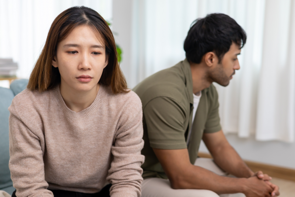 A woman sits looking down with a serious expression while a man sits behind her facing away. Both appear upset in a bright room, perhaps contemplating filing for divorce and the important questions to ask before divorce.