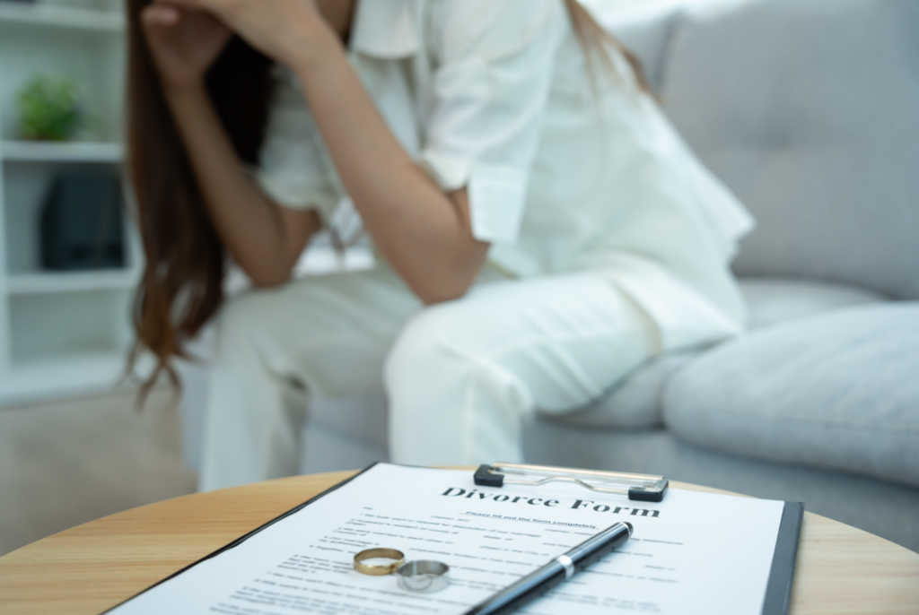 A woman sits with her head in her hands behind a table holding a California divorce form, a pen, and two wedding rings, overwhelmed by the looming divorce court deadline.