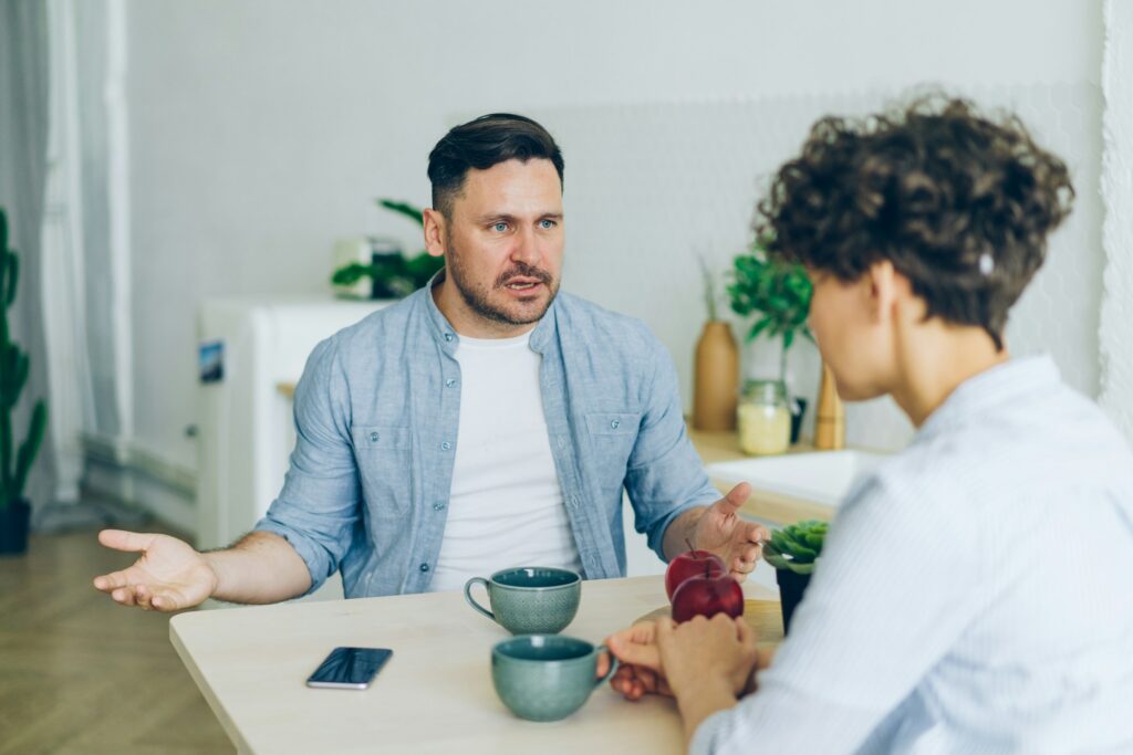 Two people sit at a table with coffee mugs, one gesturing with hands while speaking about legal separation, the other listening intently. A smartphone is on the table next to them.