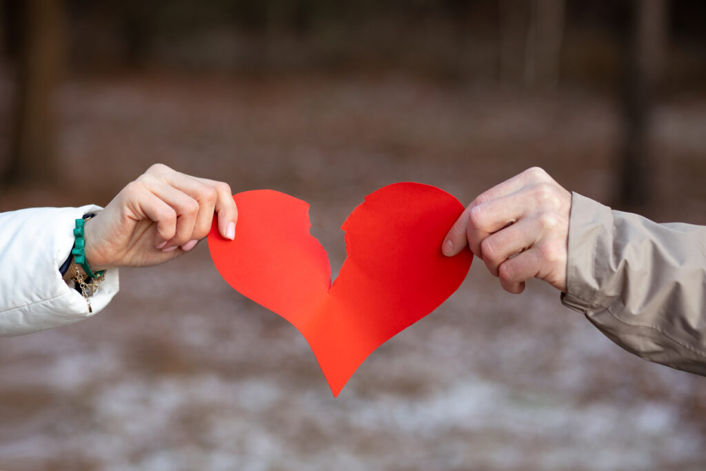 Two hands, male and female, tear red heart symbol of lovers against background of winter forest. oncept of breaking up relationship. Love is over. Resentment and separation. Negative.